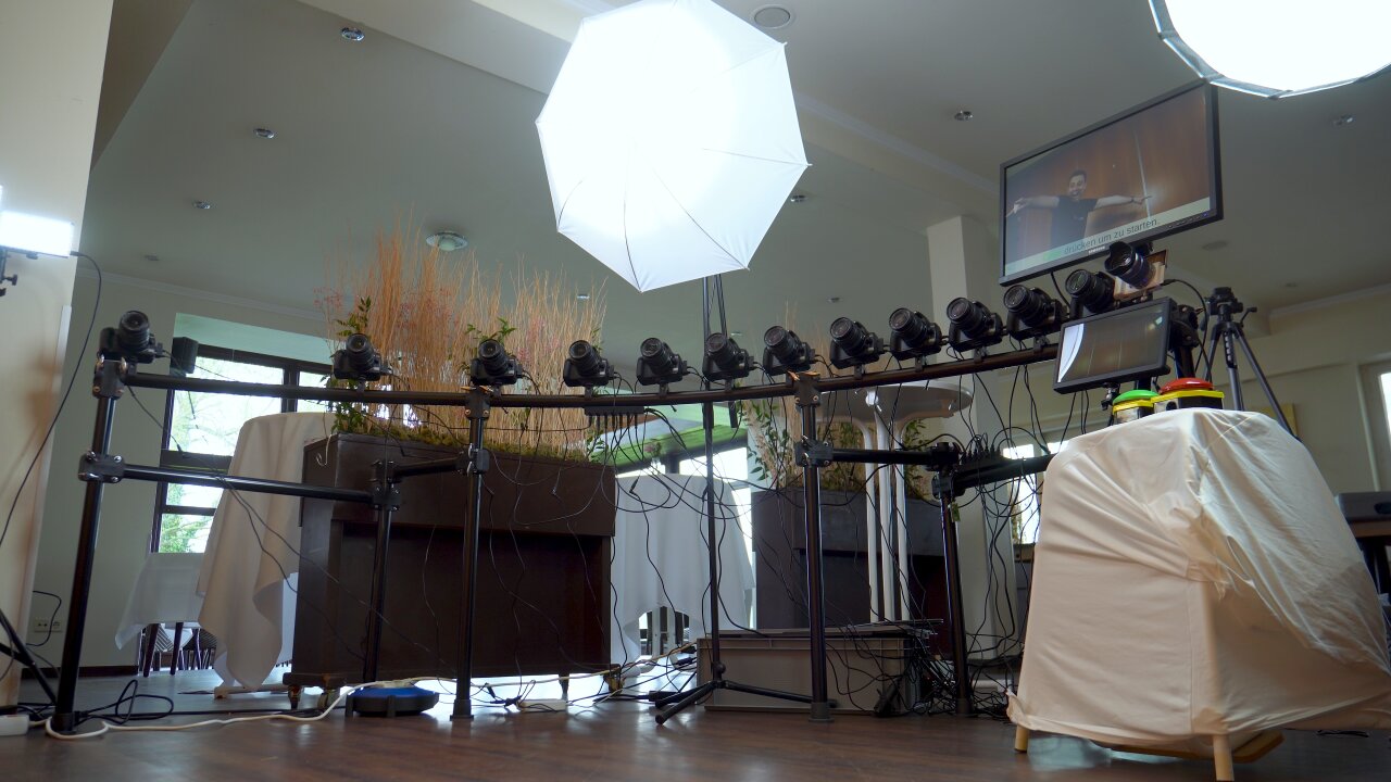 Low wide angle shot of a quarter-circle rack with twelve black and one white camera mounted on top. Bright video lights are mounted above the cameras and a room decorated for a wedding reception is visible in the background.