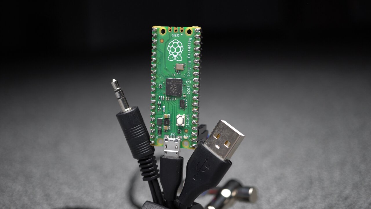 Photo of a Raspberry Pi Pico, held upright by a USB cable. The jack of an audio cable and a USB-A connector are draped next to the Pi Pico. In the back you can see several Dupont connectors attached to the Pi Pico's pins.