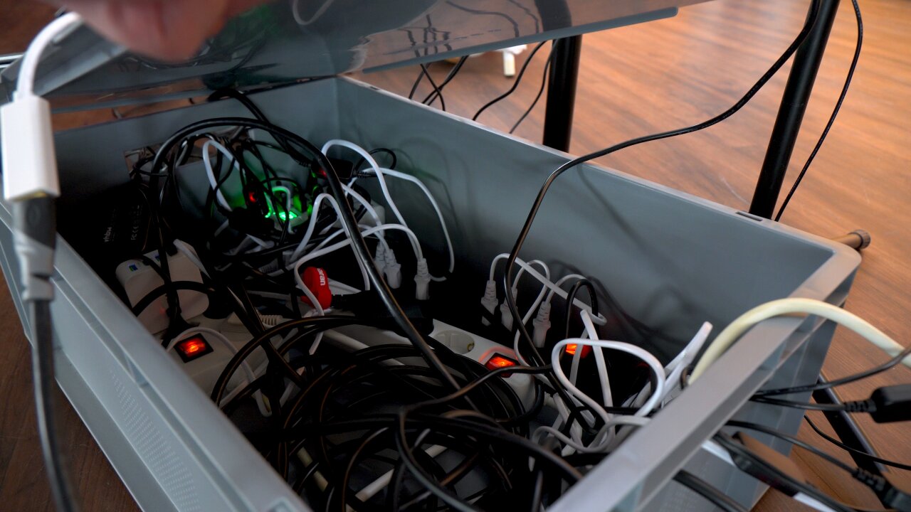 Photo of a large gray storage box with half open lid. Inside you can see many cables, power outlets, power supplies and plugs.