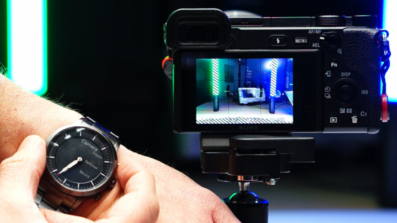 Photo of the back side of a Sony a6400 with active screen. In front of this is a wrist with a smartwatch with e-ink screen that shows the words Trigger, Selftimer and Record next to its physical buttons. A second hand is holding the watch between finger and thumb, resting the index finger on the button next to the word Trigger.