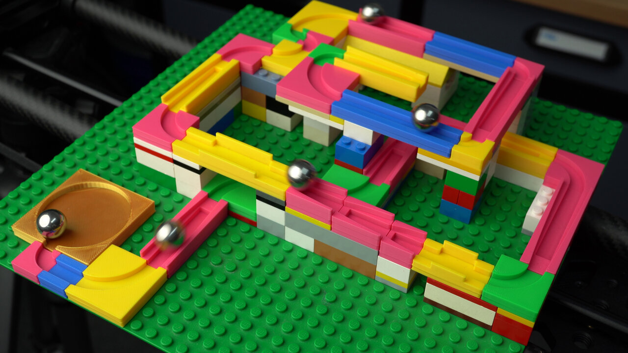 Photo of a marble run assembled from several 3D printed track pieces. They are build on a green Lego base plate with several flat Lego bricks as support. The 3D printed tracks have many different bright colors and form a complicated path with bridges. In the photo there are currently several marbles on the tracks, which are actually shiny metal ball bearings.