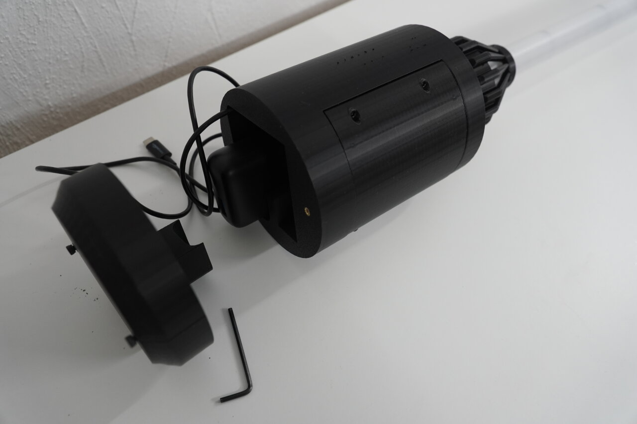 Photo of the assembled bottom part of the staff. A black cylinder with an open bottom through which the inserted power bank can be seen. The removed bottom plate lies next to it.